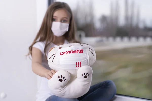 Koruyucu maskeli bir oyuncak taşıyan Coronavirus isimli küçük bir kız. Yüzünde virüsten koruyucu bir maske olan küçük bir kız elinde oyuncakla pencerenin kenarında oturuyor ve üzülüyor. Karantina.