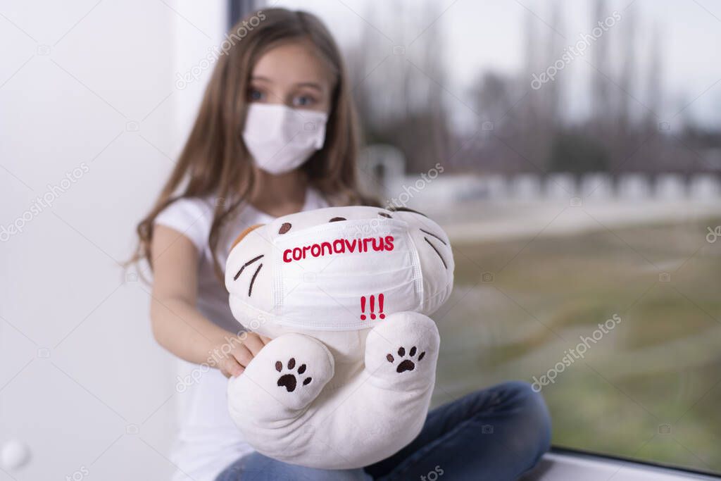 A girl with a toy in a protective mask with the text Coronavirus.A little girl t with a protective mask from the virus on her face sits by the window with a toy in her hand and is sad.Quarantine