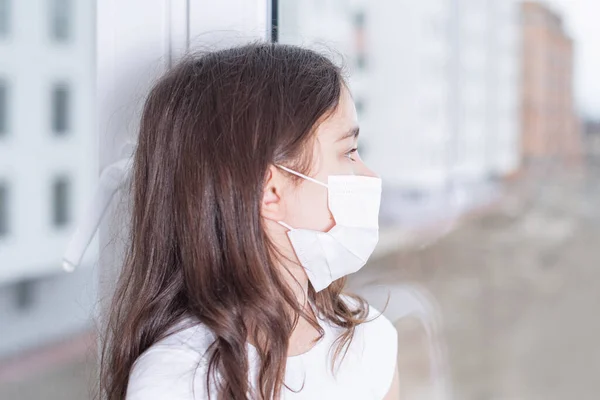 A sad girl in a protective mask from viruses on her face sits by the window.Coronavirus pneumonia COVID-19.The epidemic of coronavirus worldwide.