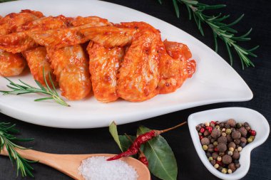 Izgara tavuk kanadı tarifi. Salamurada çiğ tavuk kanadı. Siyah arka planda baharatları olan beyaz bir fırın tabakasında marine edilmiş tavuk kanadı..