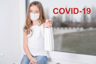 Penceredeki küçük kız yüzünde koruyucu bir maskeyle sokak koronavirüsünde. Salgın hastalık. Biyoteknoloji konsepti. Penceredeki yazı: Cauvid 19.Virüs koruma maskesi, Coronavirus pnömoni COVID-19.