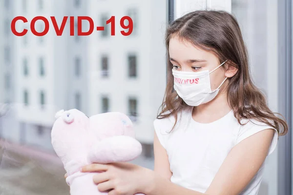 Klein meisje bij het raam met een beschermend masker op haar gezicht, op het coronavirus. Tekst op het raam covid 19.Epidemics. Begrip biotechnologie. — Stockfoto