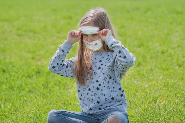 Flu epidemic, self-isolation and coronavirus protection concept.A little girl in a protective mask on her face and text coronavirus is resting on the green grass in nature.