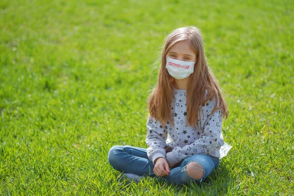 Flu epidemic, self-isolation and coronavirus protection concept.A little girl in a protective mask on her face and text coronavirus is resting on the green grass in nature.