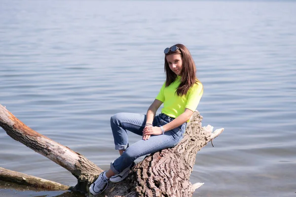 Молода приваблива дівчина з коричневим довгим волоссям, сидячи на колоді над річкою, тепло насолоджуючись сонцем . — стокове фото