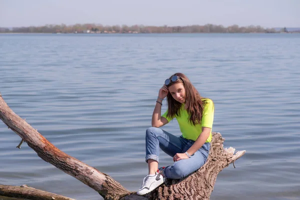 Jong aantrekkelijk meisje met bruin lang haar, zittend op een stam boven de rivier, warm genietend van de zon.Meisje op een warme lentedag. — Stockfoto