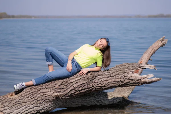 YGirl op een warme lentedag.oung aantrekkelijk meisje met bruin lang haar, zittend op een stam boven de rivier, genieten van de zon warm. — Stockfoto