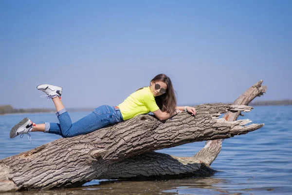 Jong aantrekkelijk meisje met bruin lang haar, zittend op een stam boven de rivier, warm genietend van de zon.Meisje op een warme lentedag. — Stockfoto
