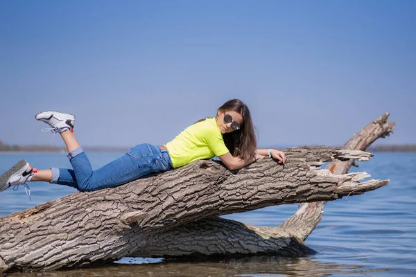 Jong aantrekkelijk meisje met bruin lang haar, zittend op een stam boven de rivier, warm genietend van de zon.Meisje op een warme lentedag. — Stockfoto