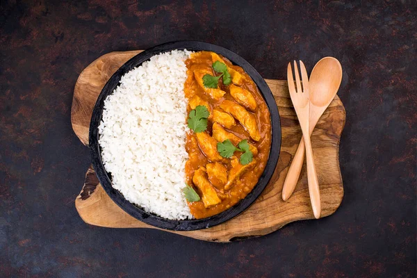 White rice with chicken and vegetables and curry sauce. Indian dish with vegetables and chicken on a black plate on a wooden kitchen board with a wooden spoon and fork.