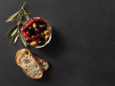 Farklı türde zeytin, kırmızı biber, ciabatta ekmeği ve koyu gri arka planda zeytin dalı içeren kabın üst görüntüsü.