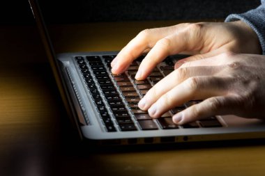 Laptopta çalışan bir adam, karanlıkta parlayan beyaz ekranla yan taraftaki ahşap bir masada görüldü.