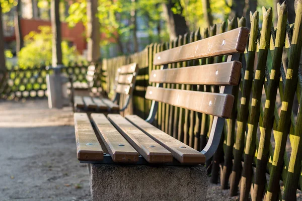A wooden bench in a park with a wooden fence behind it from the side