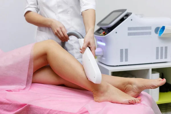 Közelkép egy női kozmetikusról orvosi köpenyben, aki egy fiatal nőt műtétként lézeres szőrteleníttet a lábával. Kozmetológia, ionizáció — Stock Fotó