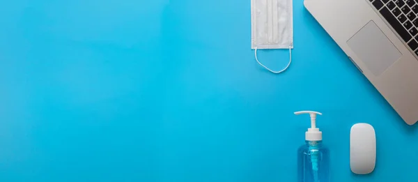 Ev kavramından çalış. Laptop, fare, tıbbi yüz maskesi mavi arka planda mavi alkollü el dezenfektanı..