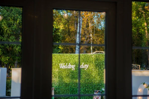 Enregistrement de la vue de la cérémonie de mariage depuis la fenêtre. Plaque en bois avec l'inscription "Jour du mariage". Fond vert. Décorations de mariage. Accessoires de mariage . — Photo