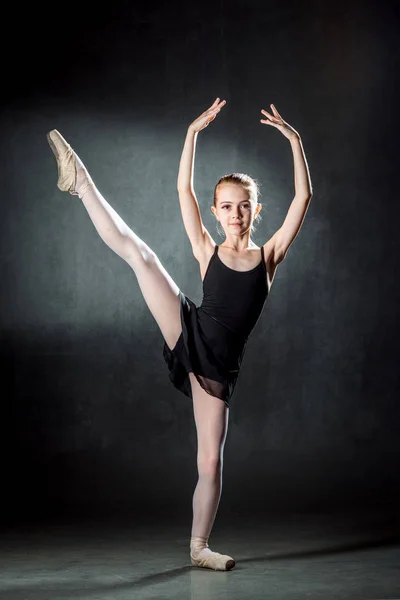 Little ballerina with hair in bunch posing to the camera. Young girl in  black costume, white tights and pointe shoes training in a ballet studio  Stock Photo