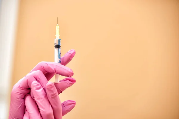 Medicinsk sprøjte i en hånd med en lyserød handske isoleret på orange baggrund. injektion - Stock-foto