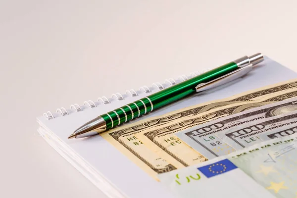 American one hundred dollar bills. Euro banknotes. A ballpoint pen and a notebook next to the bills. Business and finance. Money. White background.