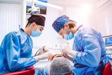 A male dentist in uniform perform dental implantation operation on a patient at dentistry office. Surgery stomatology. View from the back.