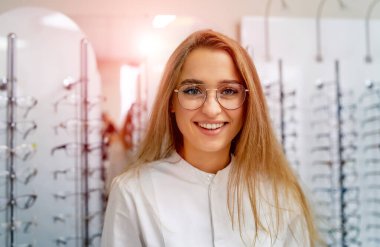 Happy female optometrist, optician is standing with raw of glasses in background in optical shop. Stand with spectacles. Eyesight correction. Blonde in spectacles.