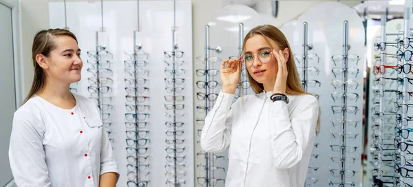 Ottico sta aiutando cliente femminile a scegliere gli occhiali in ottica. Concetto di salute degli occhi. Crudo di occhiali sullo sfondo . — Foto Stock