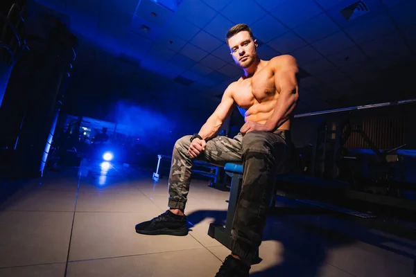 Man rests in gym after having a workout. Strong male with perfect body in the dark background with smoke. Closeup. — Stock Photo, Image