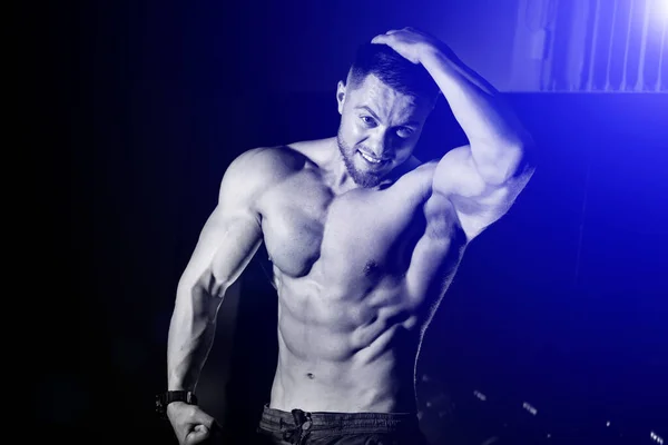 Przystojny mężczyzna z dużymi mięśniami, pozujący przed kamerą w sali gimnastycznej, czarne i niebieskie tło. Portret kulturysty z nagim tułowiem. Zbliżenie. — Zdjęcie stockowe