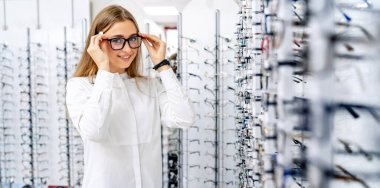 Mutlu kadın optometrist, optik dükkanında arka planda bir sürü bardakla ayakta duruyor. Optik gözlüklü kadın. Görme düzeltmesi.