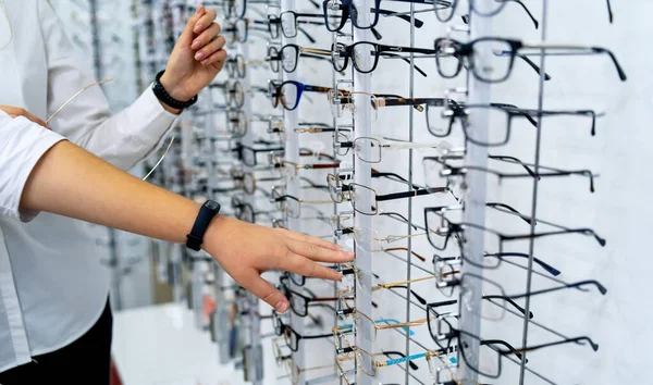 Wiersz okularów u optyków. Sklep z okularami. Stojak z okularami w sklepie optycznym. Ręka kobiety wybiera okulary. Korekta wzroku. — Zdjęcie stockowe
