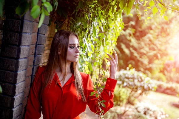 レンガの壁の近くに立って赤いドレスで魅力的な女性の写真。公園の景色。春、秋。明るい光と緑の葉の若い女の子。背景がぼやけている。閉鎖. — ストック写真