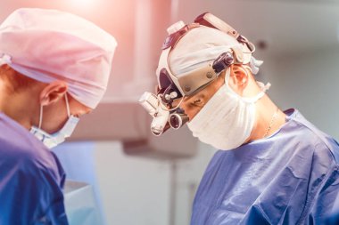 Tıbbi üniforma uzmanları hastanede bir operasyon yapıyorlar. Kafasında büyüteç olan cerrah ameliyathanede asistanlarla çalışıyor..