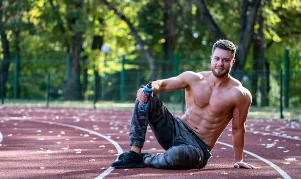Ung sportig man sitter på löparbanan och vilar efter hård träning på en stadion. Jag tittar in i kameran. Skjortlös muskulös kropp. Närbild. — Stockfoto