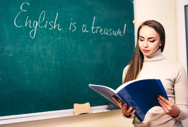Young woman with notes is standing near the blackboard, English lesson, education concept.