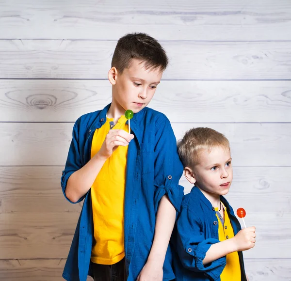 Dois brohers em camisetas amarelas e camisas azuis com pirulitos coloridos em estúdio. Adoráveis meninos estão tendo grande momento enquanto lambendo pirulitos, em um fundo de madeira leve — Fotografia de Stock