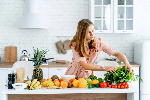 Ung och söt kvinna står i köket full av frukt och grönsaker i den moderna interiören. Livsmedel och hälso- och sjukvård — Stockfoto