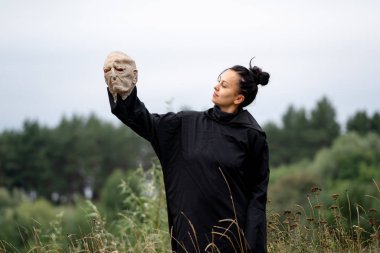 Elinde maske tutan esmer bir kadın bol siyah bir elbise ya da başlıklı bir bornoz giyiyor. Kaplama rolü açık havada.