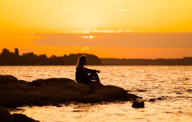 Gün batımında kadın silueti kayaya oturmuş dik dik bakıyor. Doğa ve güzellik kavramı. Turuncu gün batımı.