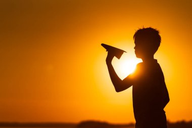 Küçük çocuk, yazın güzel turuncu gün batımında elinde bir kağıt uçak tutuyor. Gölün kenarında duran origami uçaklı bir çocuğun silueti..
