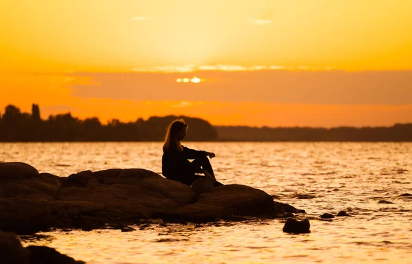 Silhuette van de vrouw bij zonsondergang, zittend op een rots die er recht uitziet. Natuur en schoonheid concept. Oranje zonsondergang. — Stockfoto