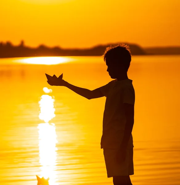 A fiú papírhajót tart a víz ellen. Gyönyörű narancssárga naplemente. Papírhajó. Origami vagyok. Narancssárga napnyugta. Boldog gyermekkori koncepció — Stock Fotó