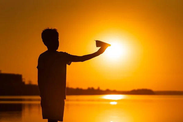 El niño sostiene un avión de papel en la mano al atardecer. Un niño levantó la mano al cielo y juega con el origami por la noche en el lago. Silhuette. — Foto de Stock