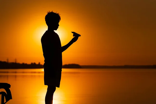 Teenie-Junge betrachtet Papierflugzeug in der Hand bei orangefarbenem Sonnenuntergang. eine Silhouette eines Kindes mit Origami-Flugzeug gegen Wasser. — Stockfoto