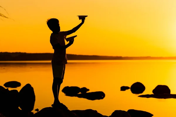 Küçük çocuk elinde iki kağıt uçak tutuyor ve gün batımında gölün arka planında oynuyor. Gün batımına karşı Siyah Siluet — Stok fotoğraf