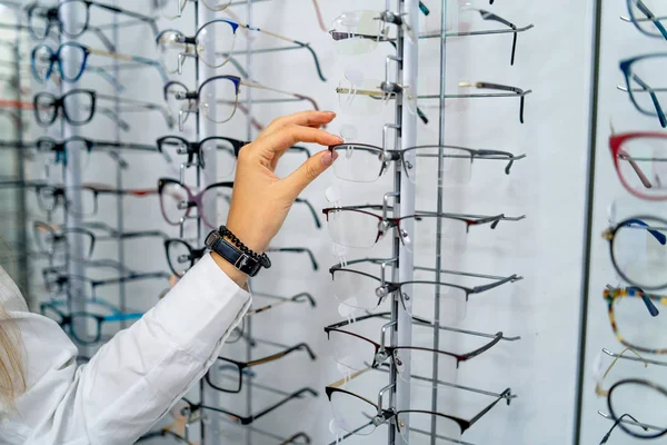 ऑप्टिशियनवर चष्माची पंक्ती. चष्मा दुकान. ऑप्टिक्सच्या स्टोअरमध्ये चष्मासह उभे रहा. महिला हात चष्मा निवडून. चष्मा सादर करणे . — स्टॉक फोटो, इमेज