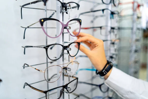 Fila de gafas en una óptica. Tienda de anteojos. Soporte con gafas en la tienda de óptica. Las mujeres eligen gafas a mano. Presentación de espectáculos . — Foto de Stock