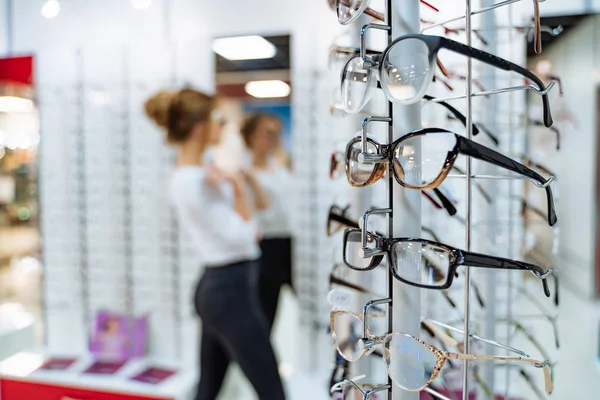 光学系の店内にメガネで立ってください。女性は背景のぼやけたガラスを選択します. — ストック写真