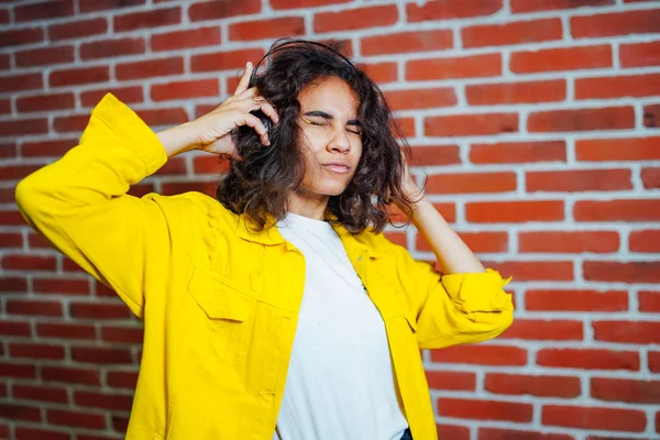 Studio portrait of grumpy young female wearing yellow sweater, with headphones on head, unssatisfied with bad sound music, on brick wall background. Closeup