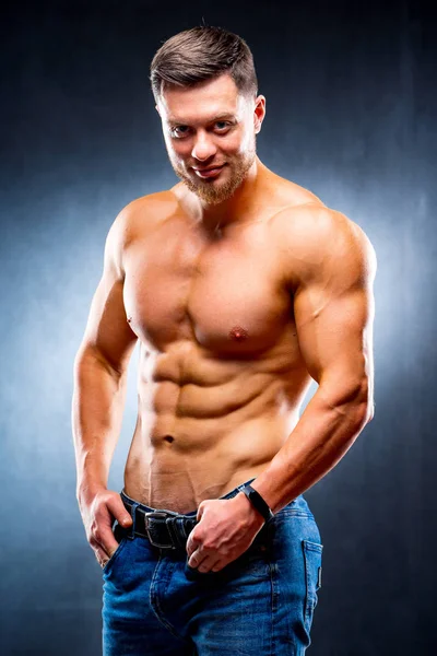 Sexy young man on a dark background posing. Perfect male body. Hands in pockets. Male beauty concept. Half turned to the camera. Closeup. — Stock Photo, Image