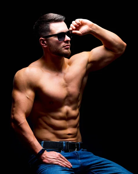 Pues construye un hombre guapo sin una camisa posando en el estudio. Cuerpo fuerte y cara fría. Hombre con gafas, sentado en una silla con las manos en las piernas. enfoque selectivo. Primer plano. . —  Fotos de Stock